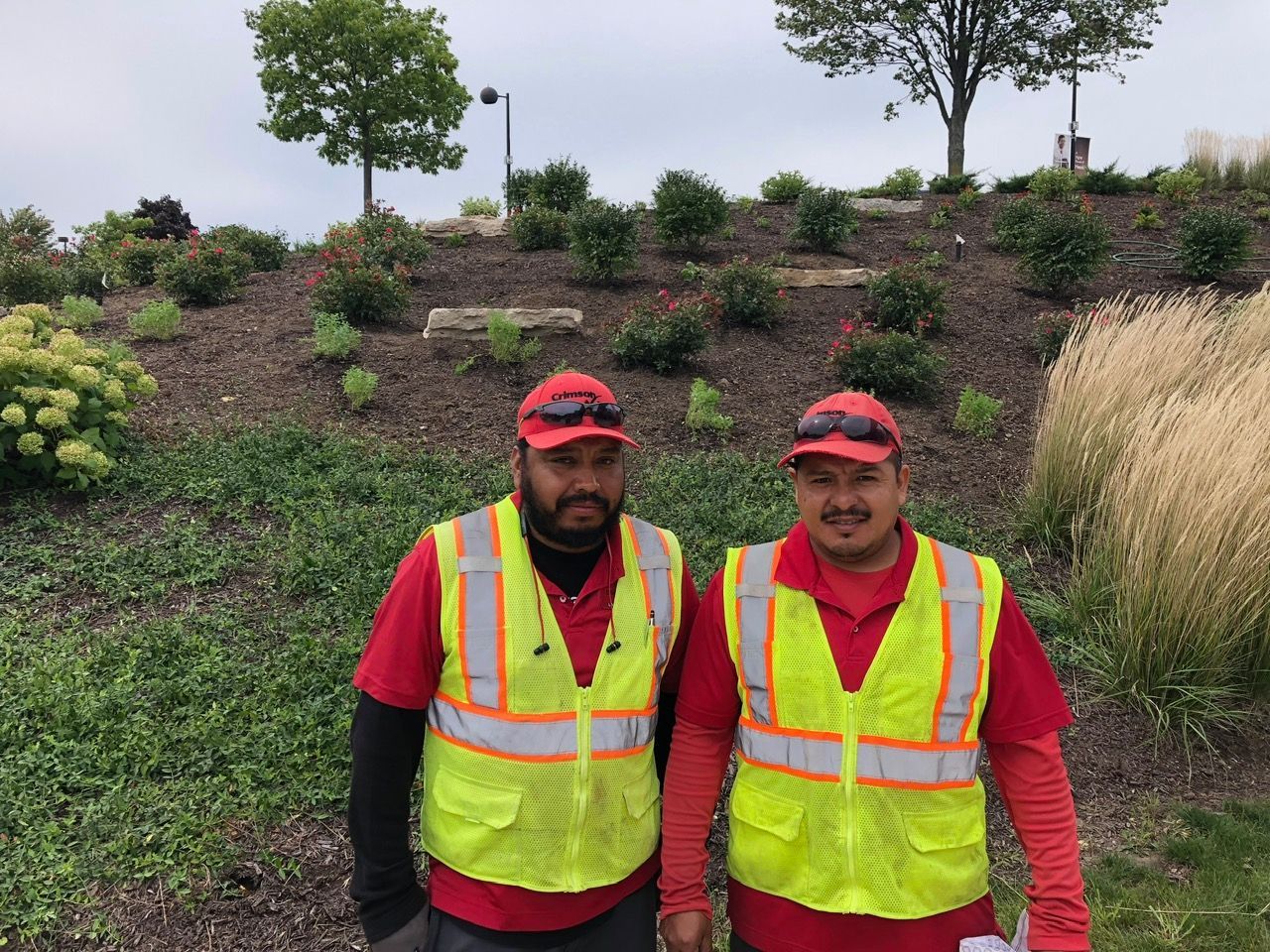 Two workers in red shirts and reflective vests with matching red caps stand in front of a landscaped garden.