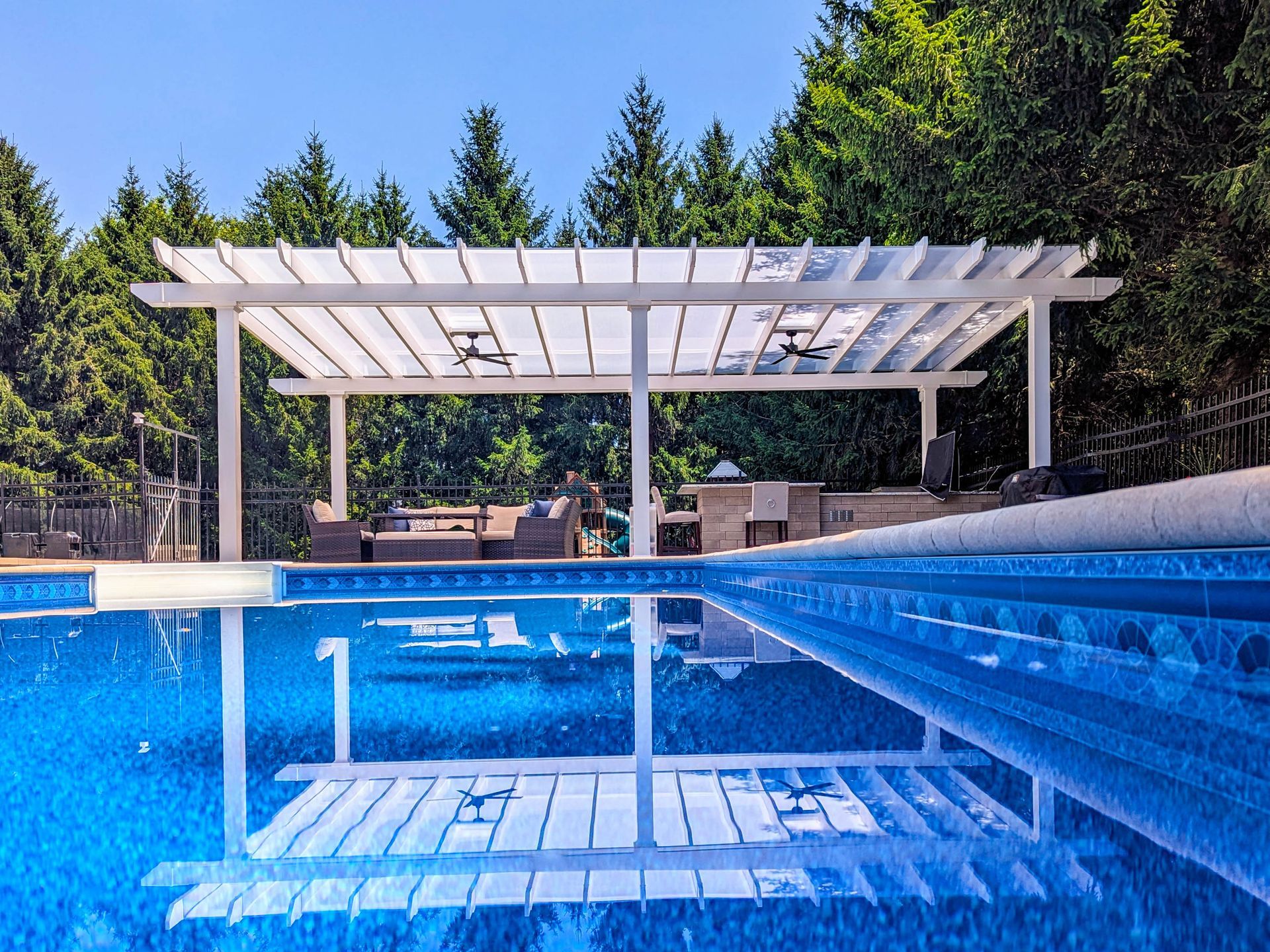 A white pergola sits beside a bright blue swimming pool, reflecting on the water surface with trees in the background.