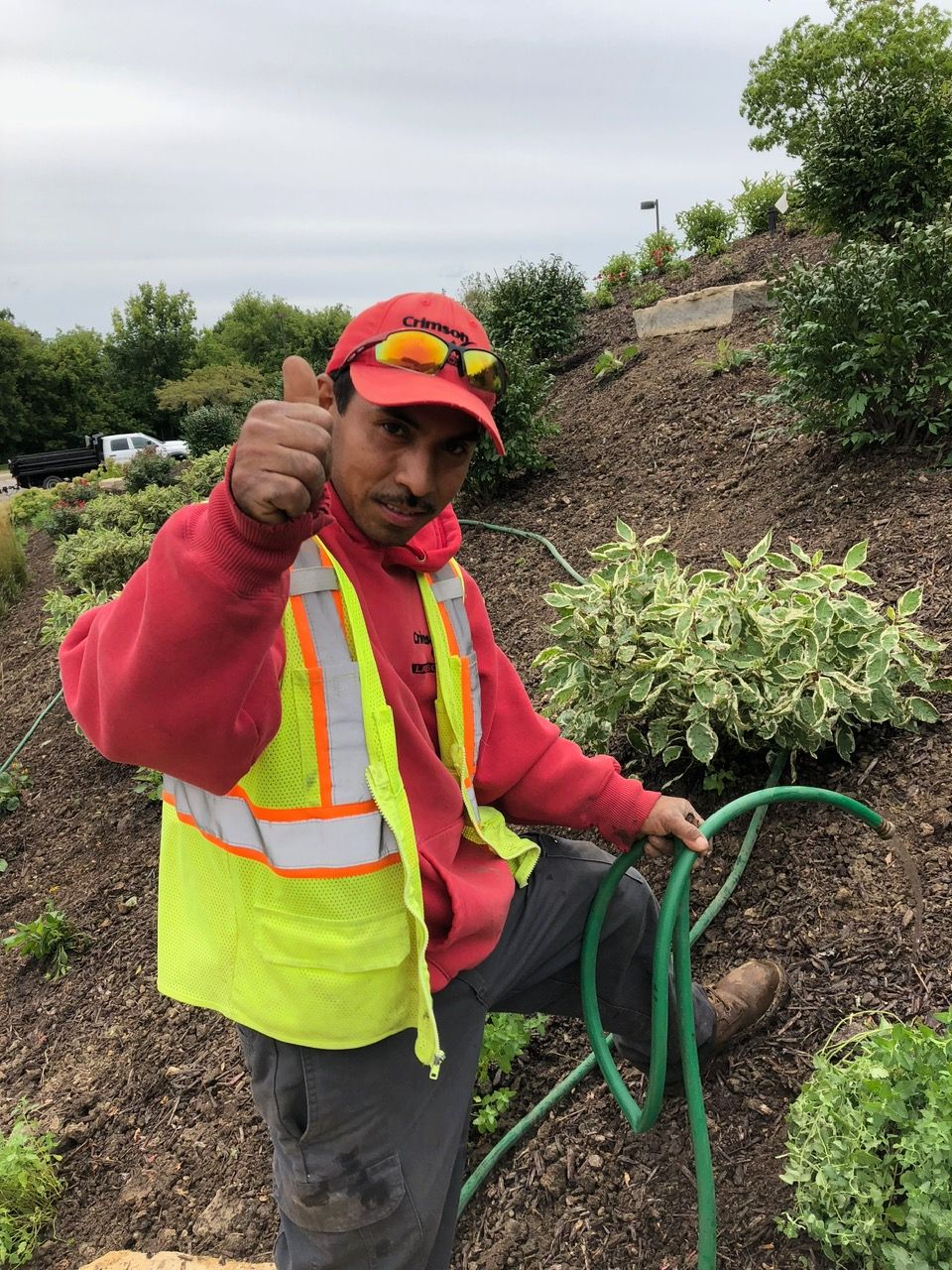 A worker in a red hoodie and high-visibility vest gives a thumbs-up while holding a green garden hose on a mulched hill.