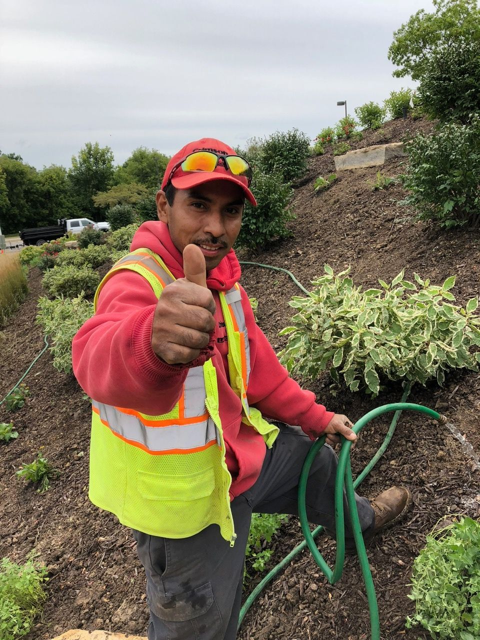 A worker in a red hoodie and neon safety vest gives a thumbs up while holding a green garden hose in a landscaped area.