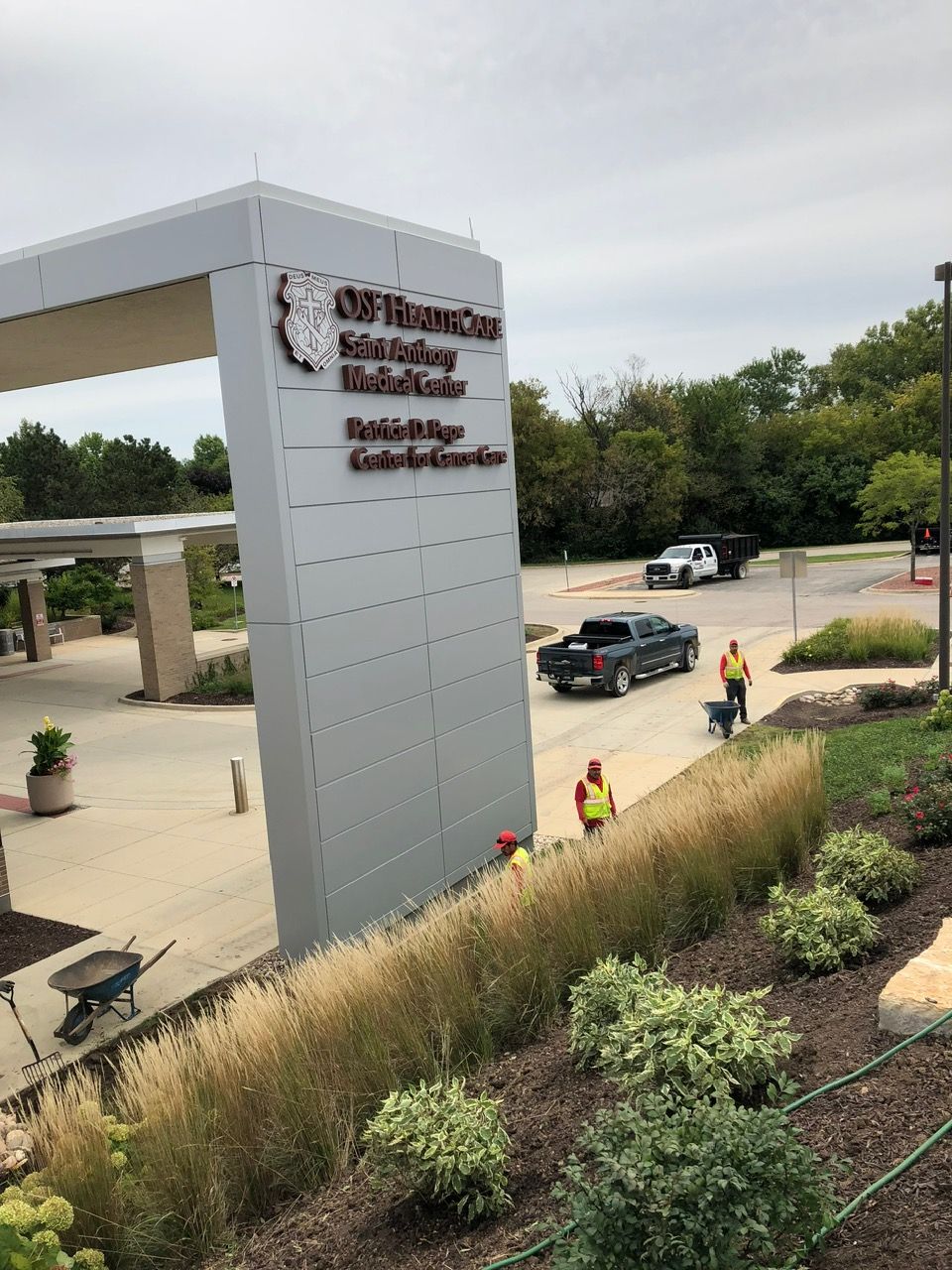 A tall, grey sign for Advocate Schaumburg Medical Center stands outside with workers in safety vests nearby.