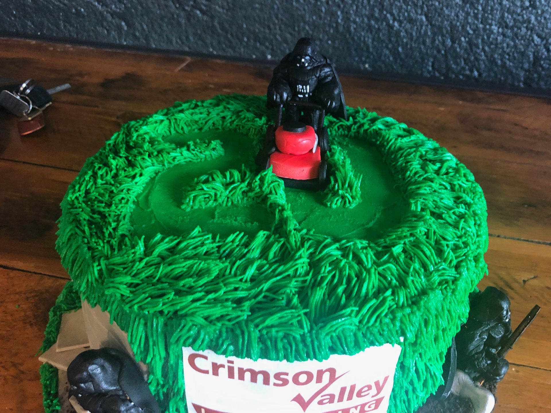 A green frosted cake featuring a miniature figure mowing a grass-like top, with figurines on the side and a logo on front.