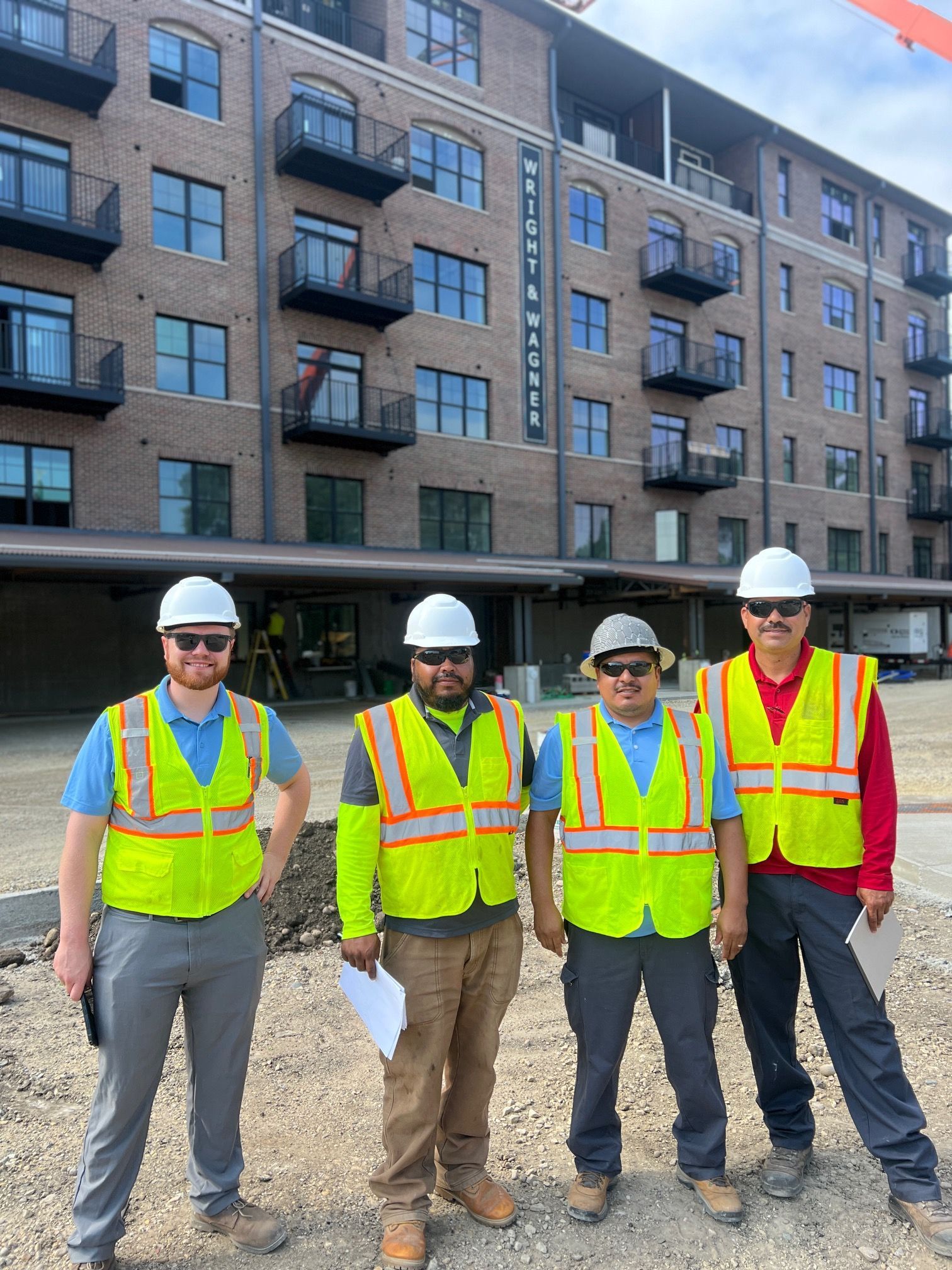 Four construction workers in safety vests and hard hats stand outside a multi-story brick apartment building.