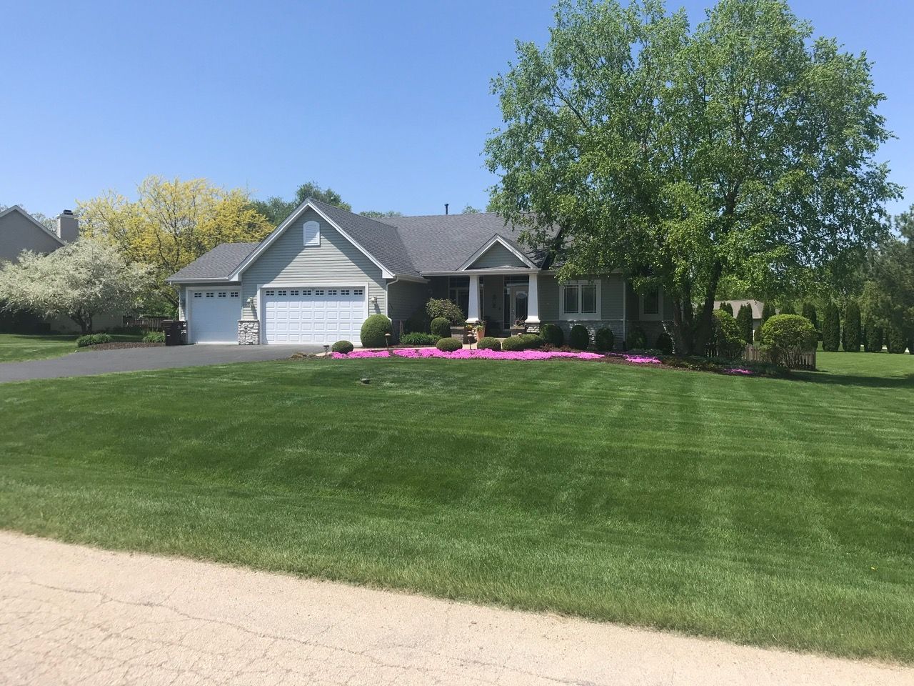 A light gray house with a white garage and front columns, surrounded by a large green lawn, trees, and bright pink bushes.