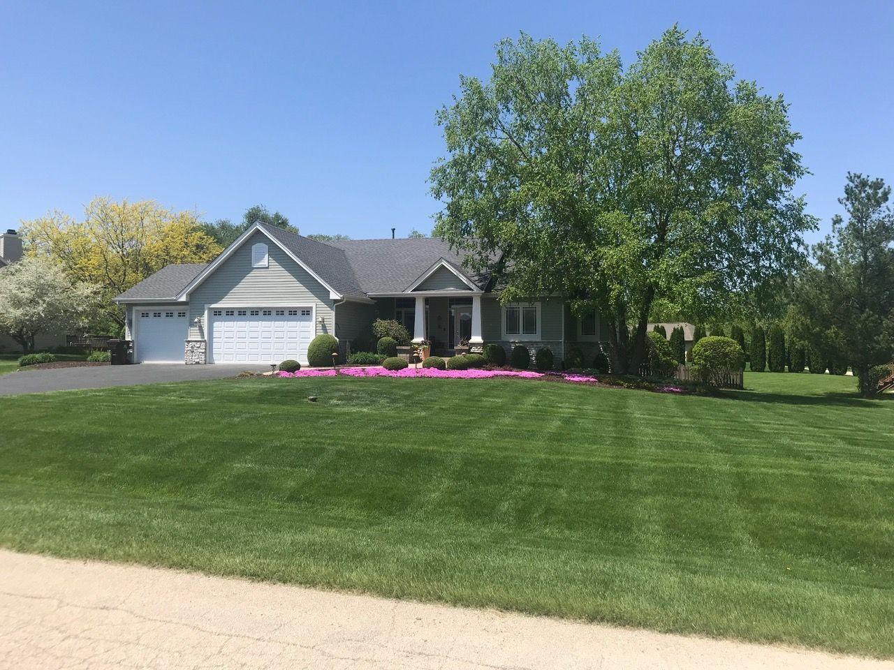 A gray suburban house with a white garage and a large, well-manicured lawn under a clear blue sky.