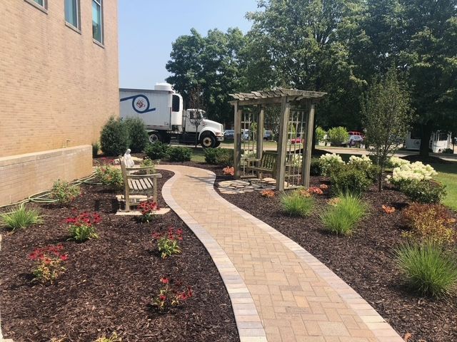 A curving brick walkway leads past a wooden pergola and a garden bench next to a brick building on a sunny day.