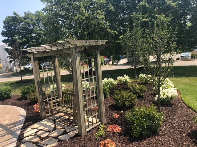 A wooden garden arbor with a bench sits on stone pavers surrounded by mulch, green shrubs, and white flowering bushes.