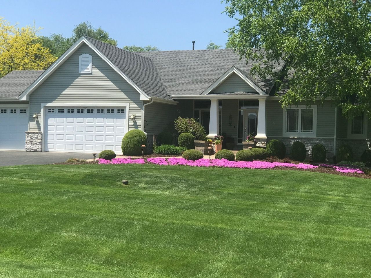 A single-story suburban house with a two-car garage, gray siding, and a large front lawn bordered by pink flowers.