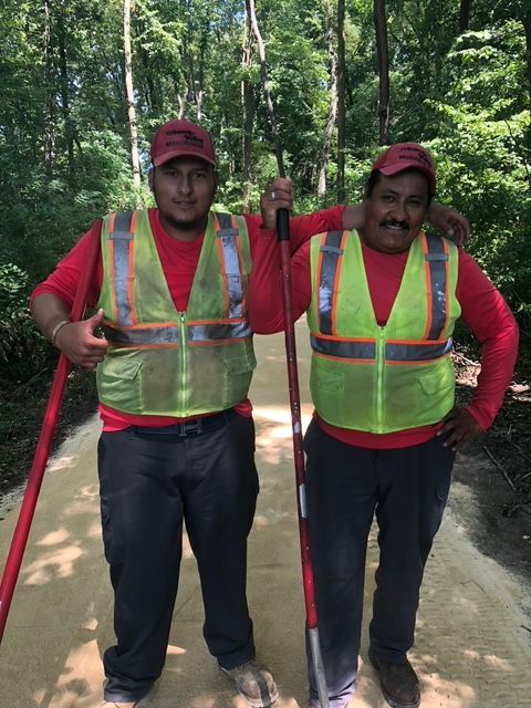 Two workers in red long-sleeve shirts and safety vests stand on a gravel path in a forest, holding tools and smiling.