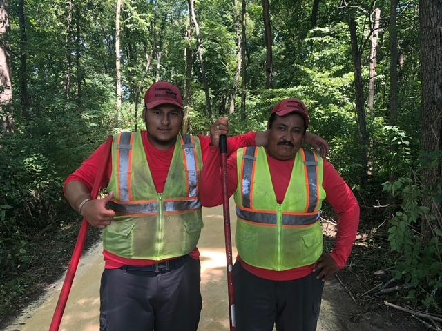 Two workers in red shirts and neon safety vests standing on a wooded trail, each holding a long-handled tool.