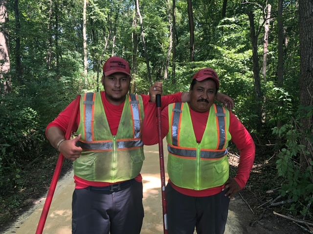 Two workers in red shirts and reflective vests pose on a wooded path, holding long-handled tools in a forest setting.