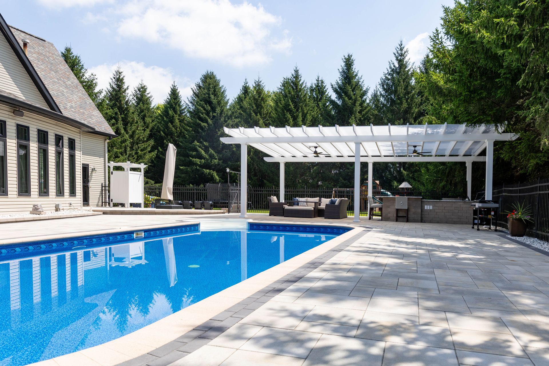 A sunny backyard featuring a rectangular swimming pool next to a stone patio with a white pergola and outdoor seating.