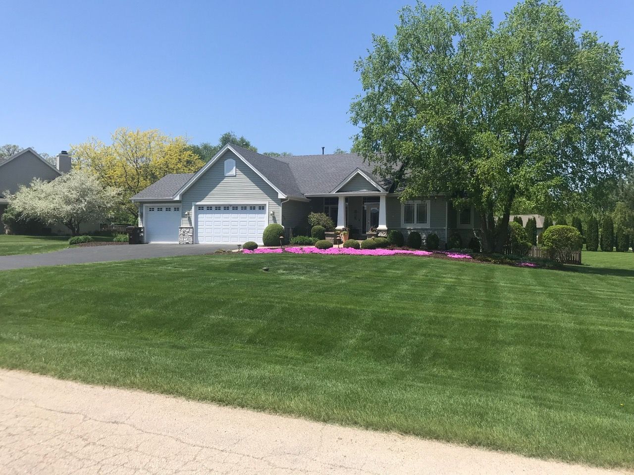 A single-story suburban house with a two-car garage, manicured lawn, and blooming pink flowers in front.