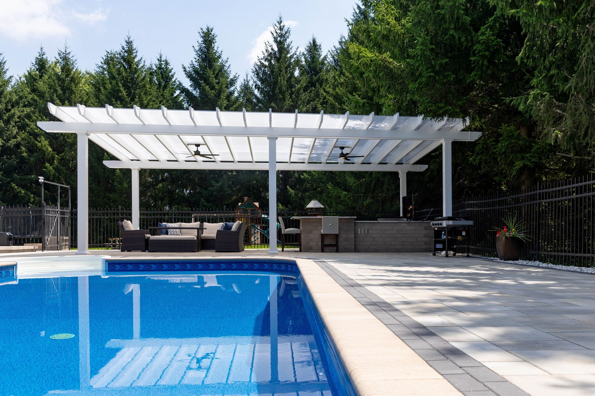 A blue backyard pool next to a white pergola covering a patio with outdoor seating and a grill area.
