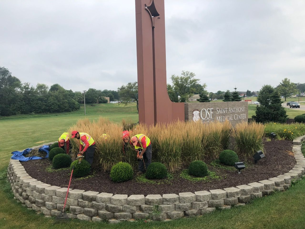 Three workers in high-visibility vests prune ornamental grasses in a landscaped stone planter near a tall monument sign.