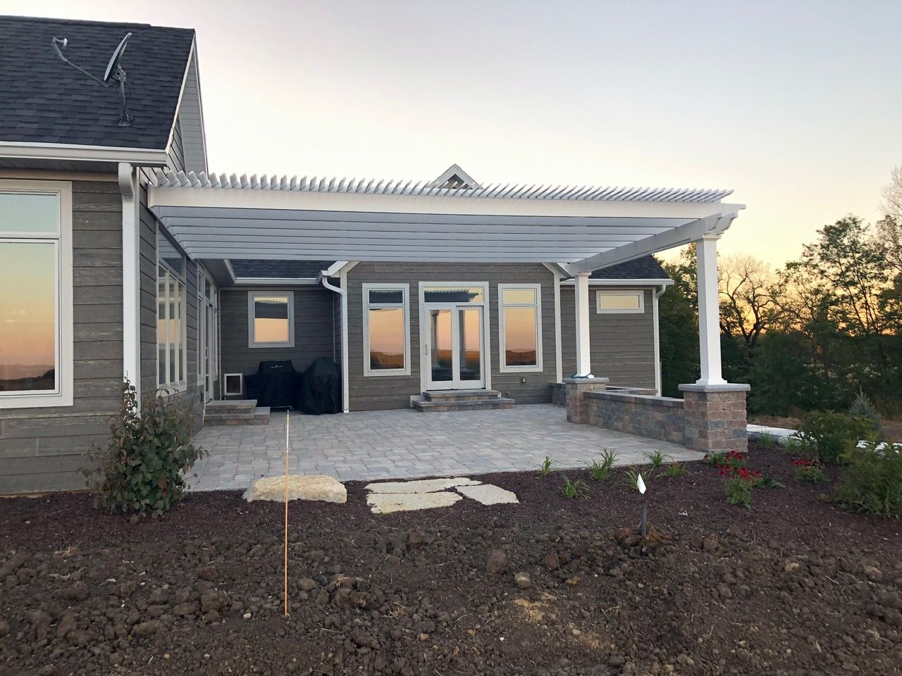 Gray house with a paved patio, white pergola, stone seating, and a dirt yard at sunset.
