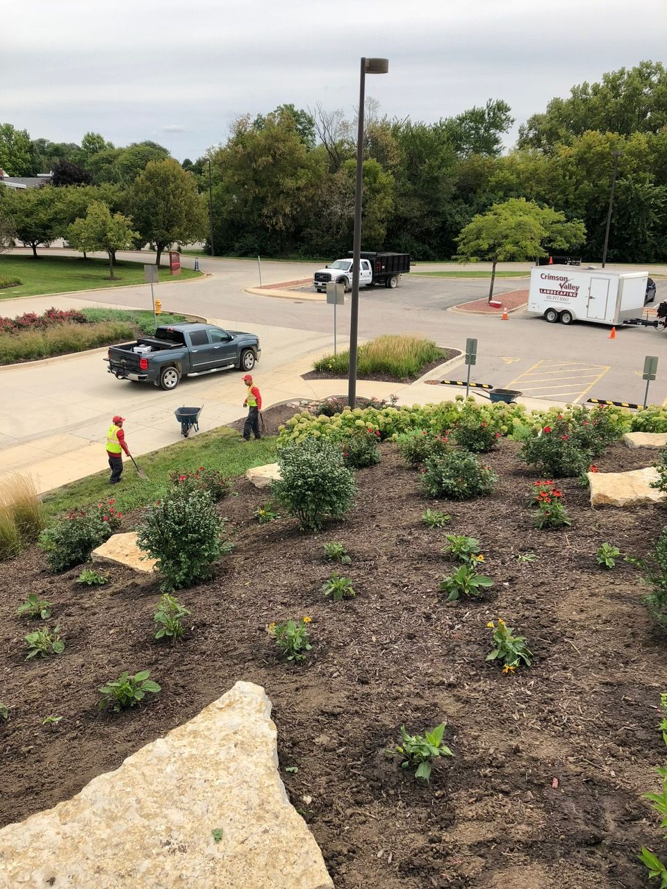 Two workers in safety vests tend to a garden slope with stone accents, while trucks and a utility trailer park nearby.