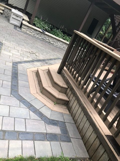 A ground-level view of a patio with light gray pavers, dark stone accents, and wooden deck steps leading up to a railing.