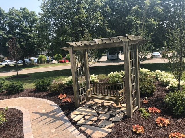 A wooden garden arbor with a swing bench, surrounded by landscaped flower beds and a paved stone path in a park.