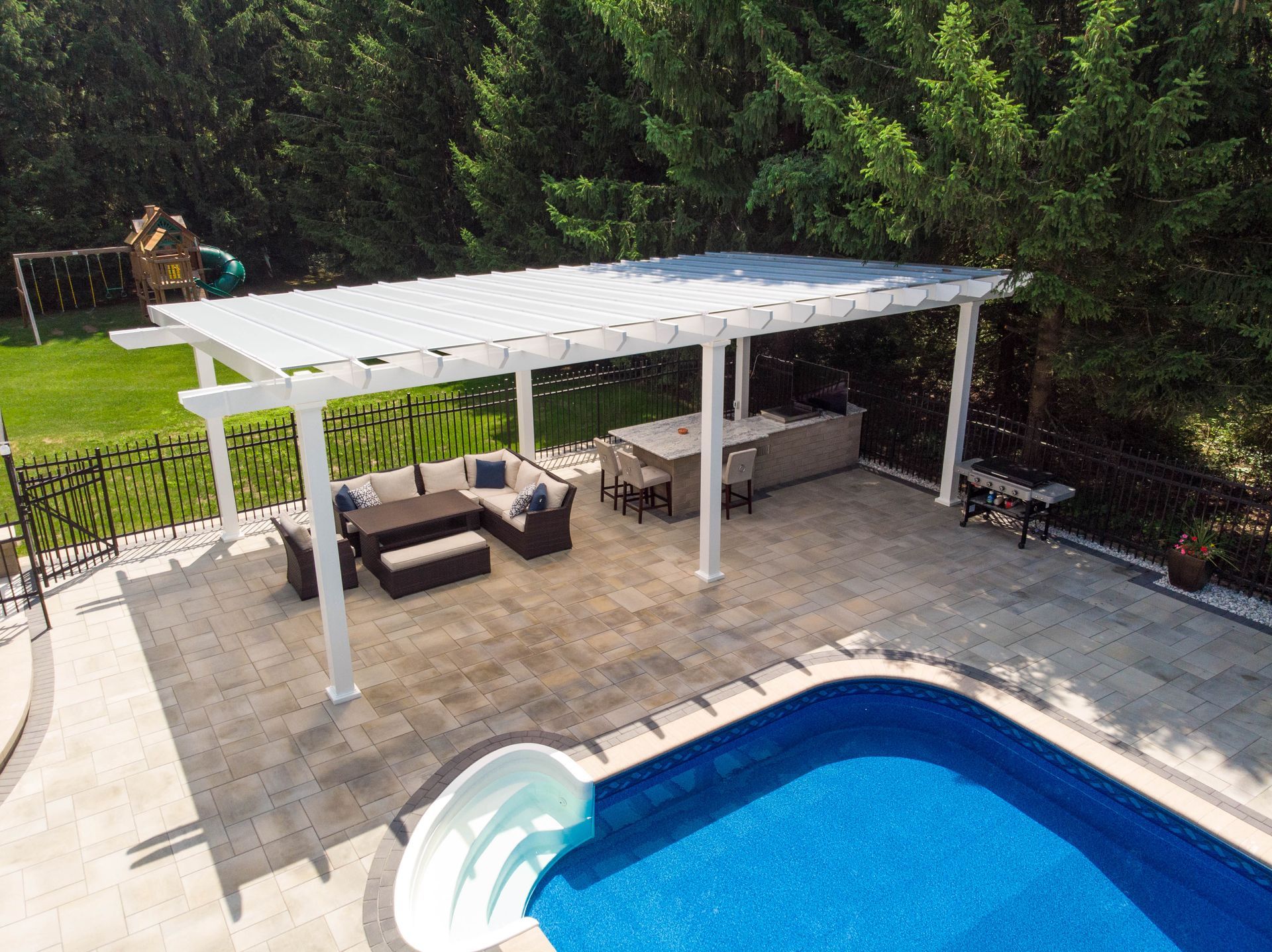 An aerial view of a white pergola over a patio with outdoor seating, a kitchen island, and an adjacent swimming pool.