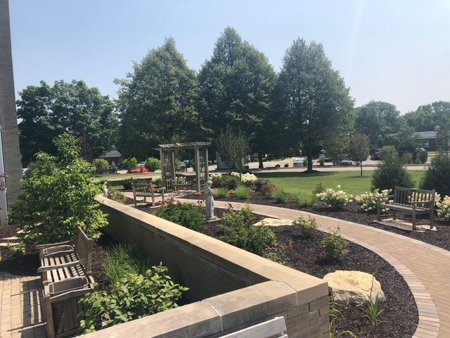 A landscaped garden featuring a curved brick path, stone retaining wall, wooden benches, and a small pergola under trees.