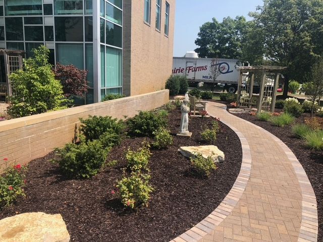 A curved brick walkway leads past a garden bed with shrubs, mulch, a stone statue, and a building with large windows.