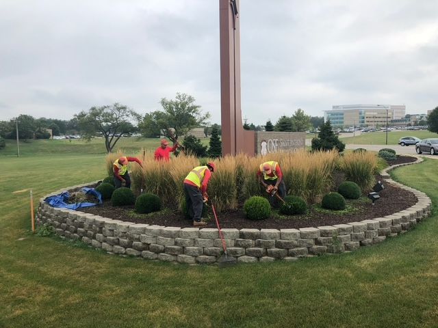 Four people in high-visibility safety vests working in a landscaped garden bed with tall grass and shrubs around a sign.