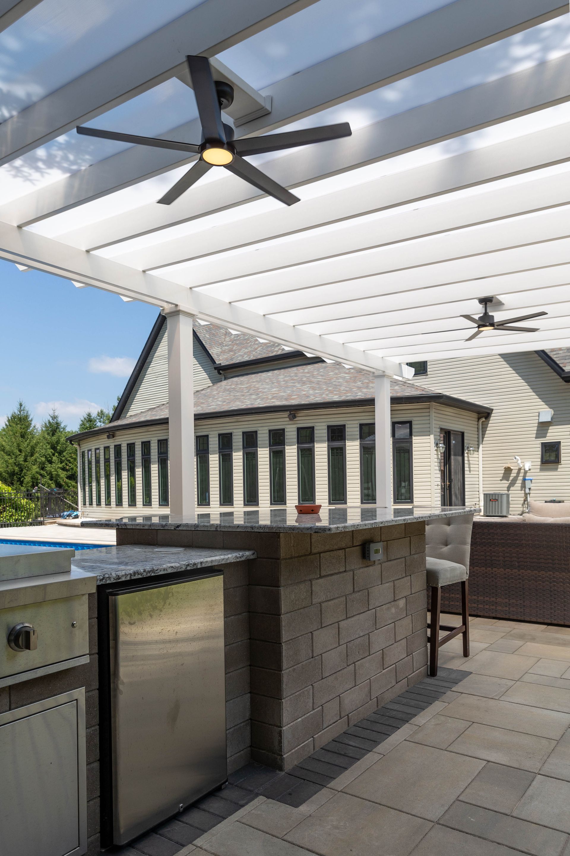 A modern outdoor kitchen with a stone bar, stainless steel grill, and refrigerator under a white wooden pergola.
