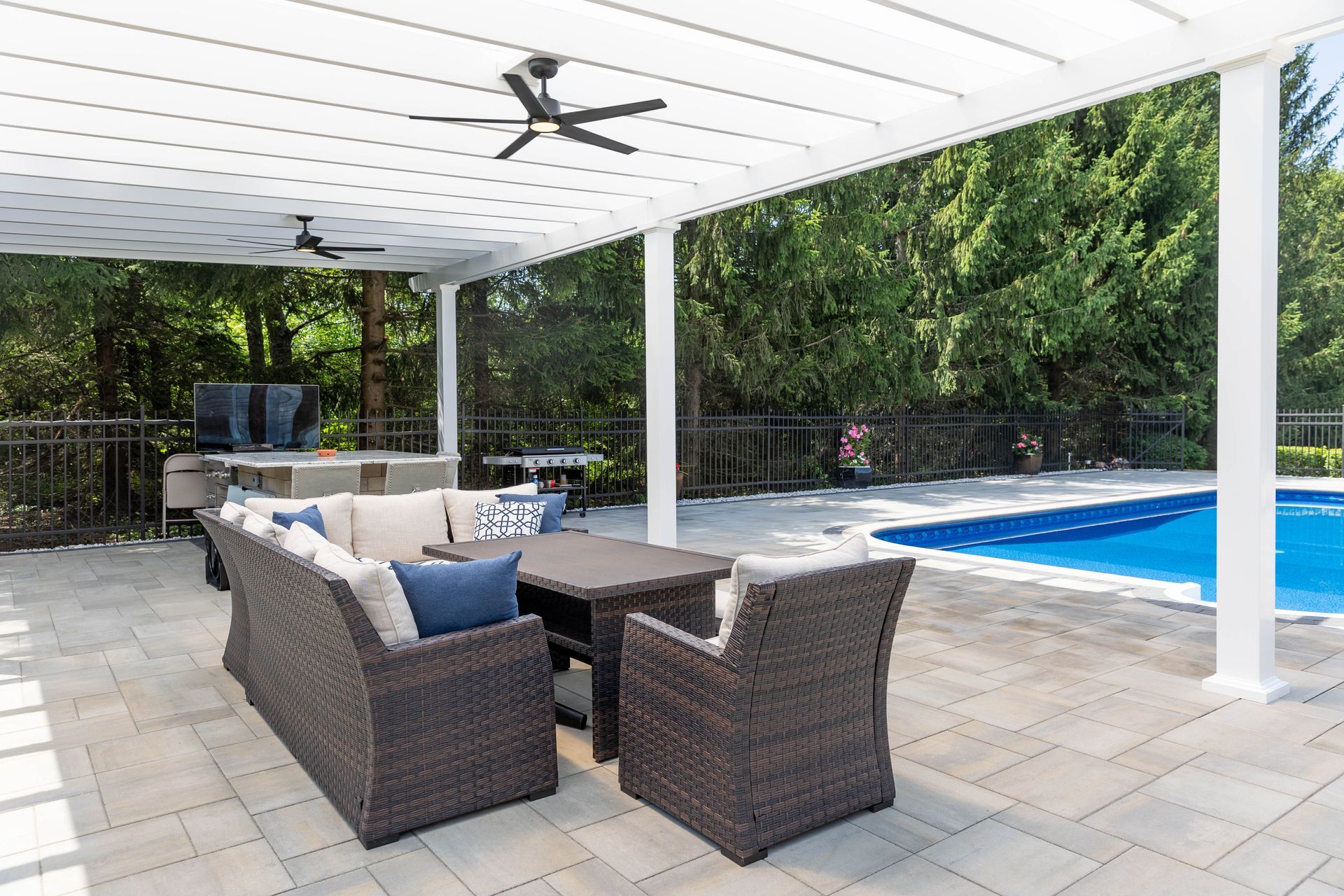 A patio with a brown wicker sofa and chairs under a white pergola next to a swimming pool, surrounded by trees.