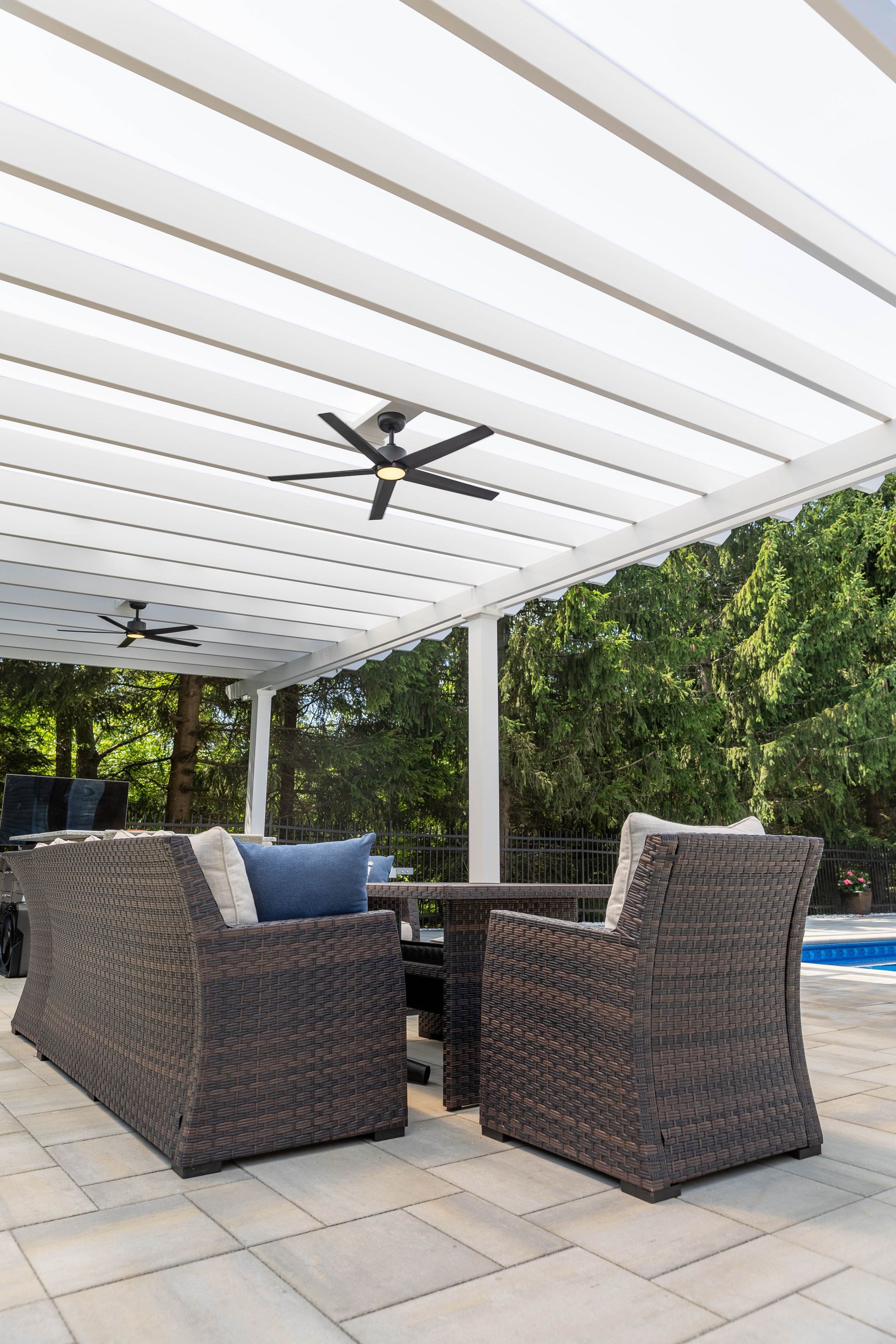 Outdoor wicker patio furniture under a white pergola with ceiling fans, next to a pool area with trees.