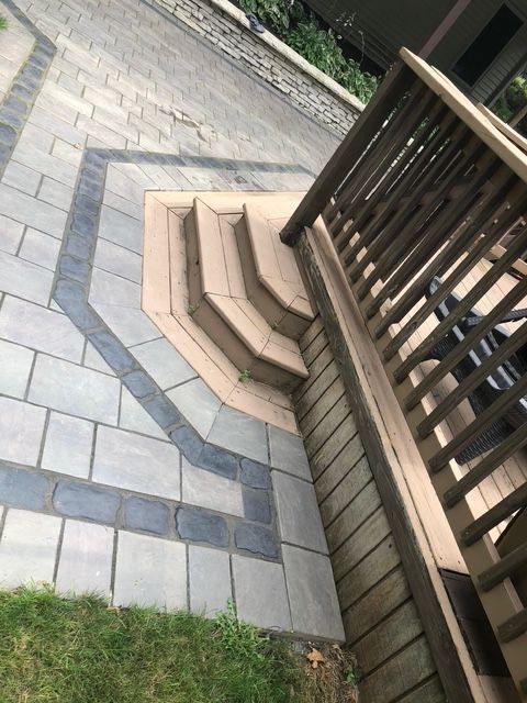A wooden deck with a small set of stairs leading down to a light-colored patio featuring a dark grey stone border.