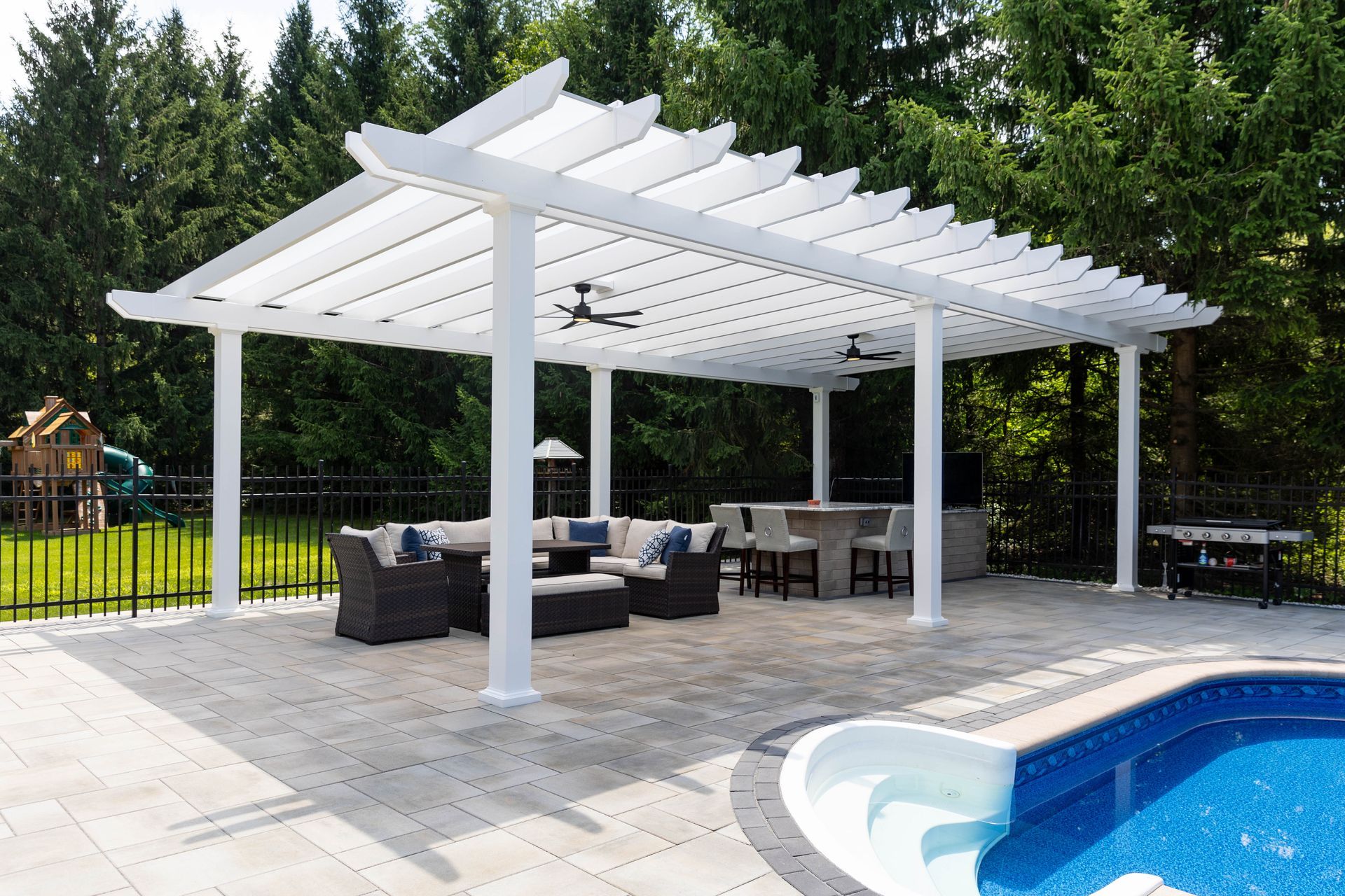 A white pergola over a patio seating area with outdoor furniture and fans, located next to a swimming pool.