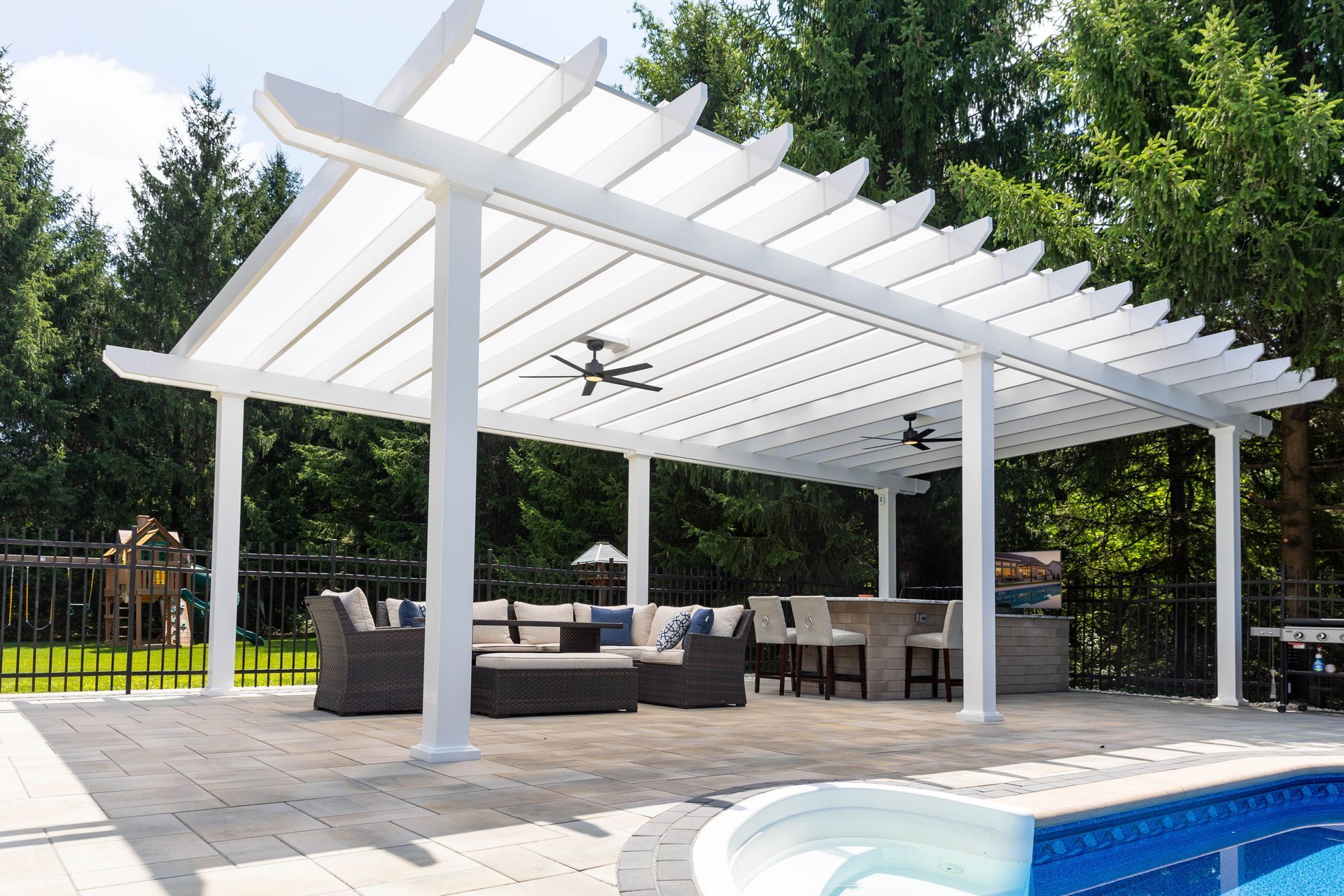 White pergola over a patio with outdoor sofa seating and a bar area by a pool, set against a background of green trees.