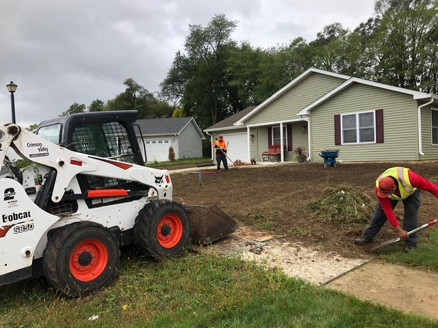 A white skid-steer loader and two workers in high-visibility vests work on landscaping a residential front yard.
