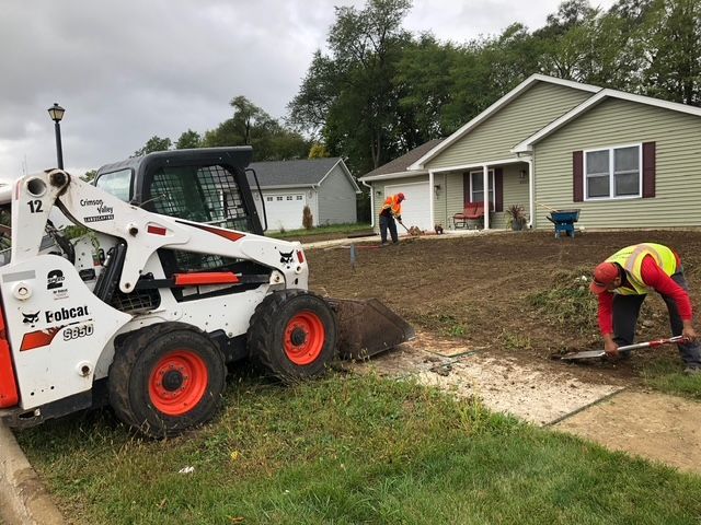 Workers use a white Bobcat skid steer and hand tools to clear and level the lawn in front of a house.