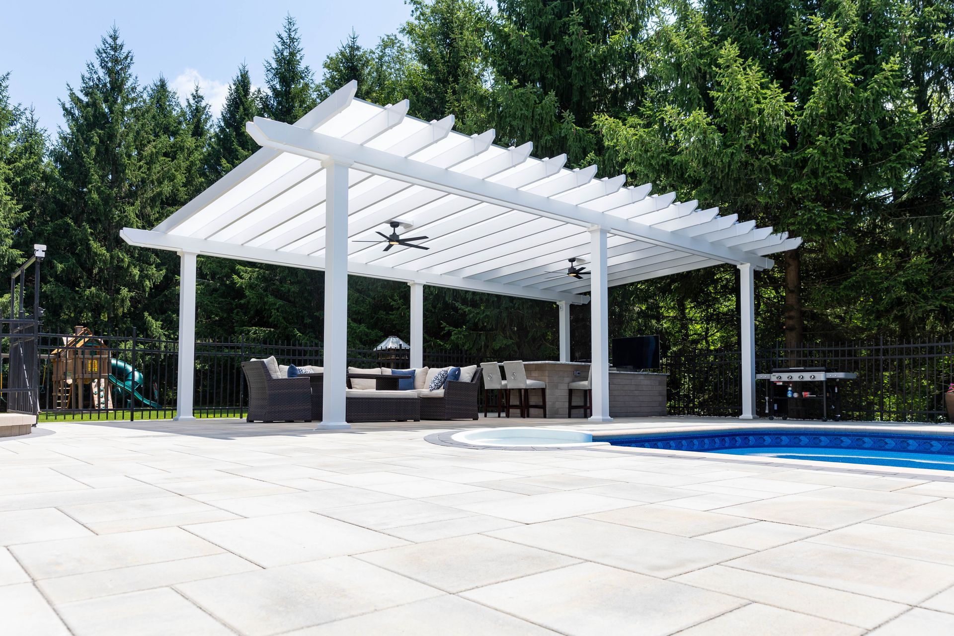 White pergola covering a patio lounge area with furniture next to a swimming pool in a backyard.