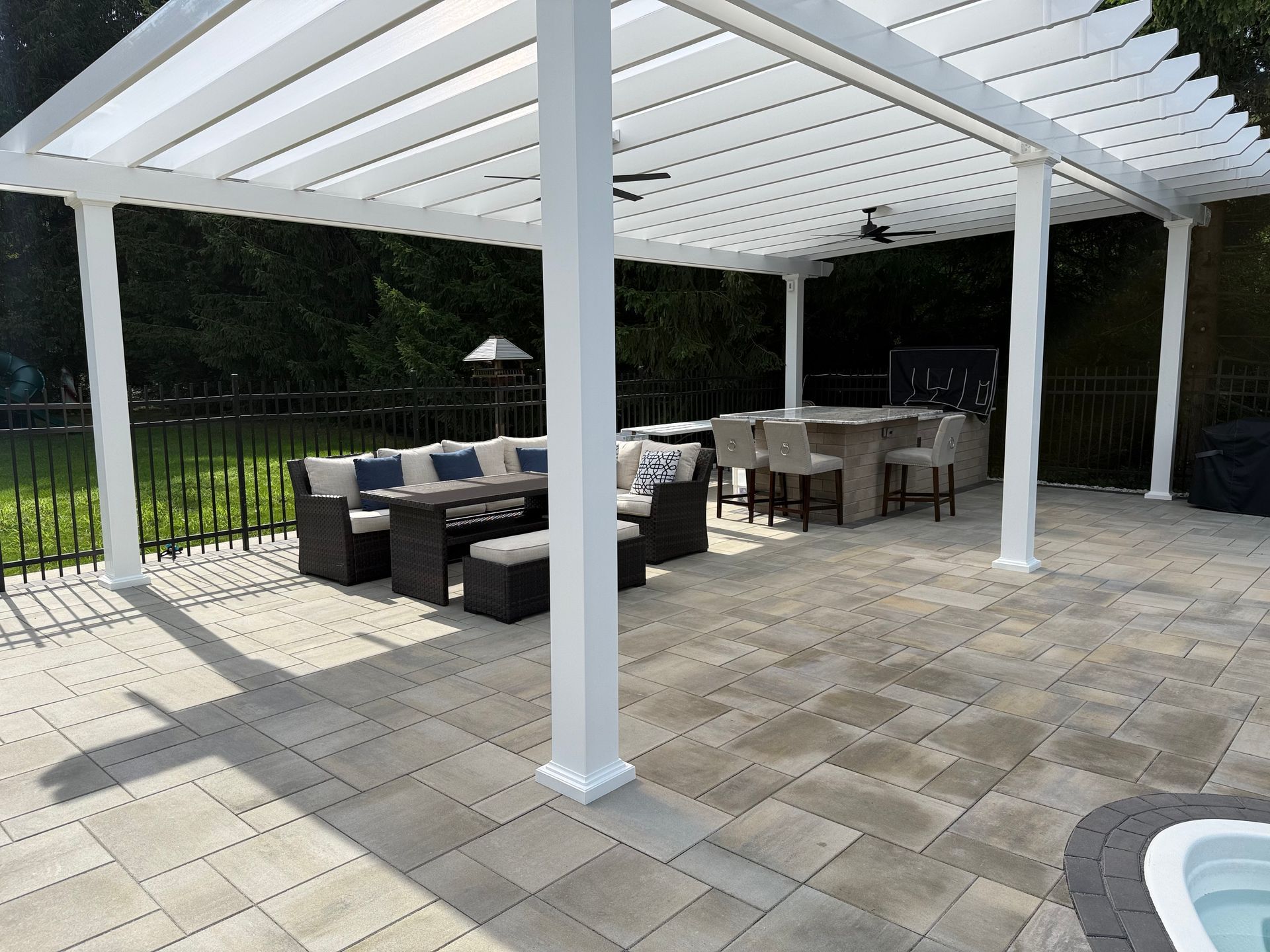 A white pergola covers a stone patio with outdoor lounge seating and a stone bar area in a backyard setting.