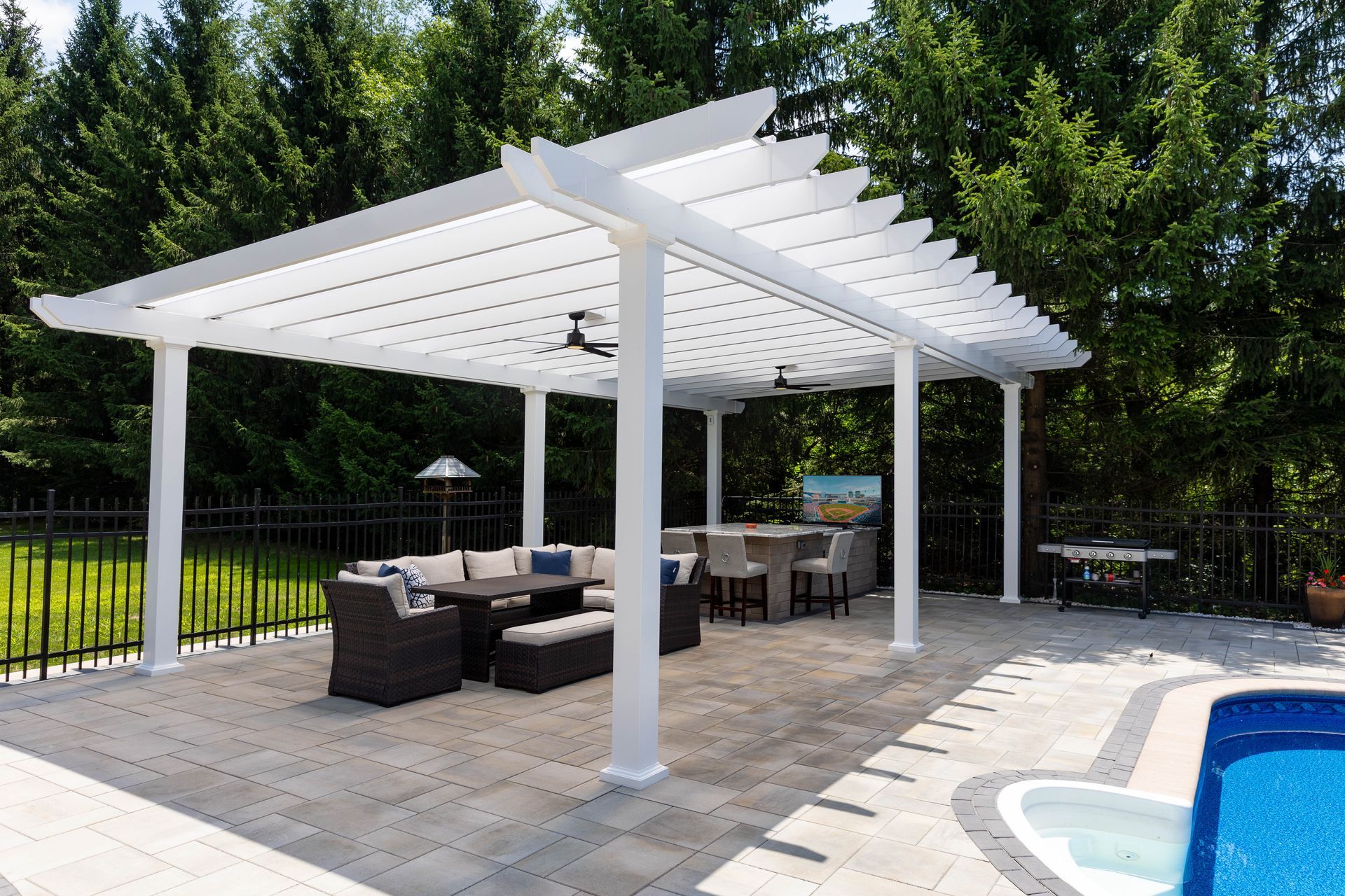 A white pergola over a patio with outdoor sofa seating, a high-top table, and a pool, set against a backdrop of trees.