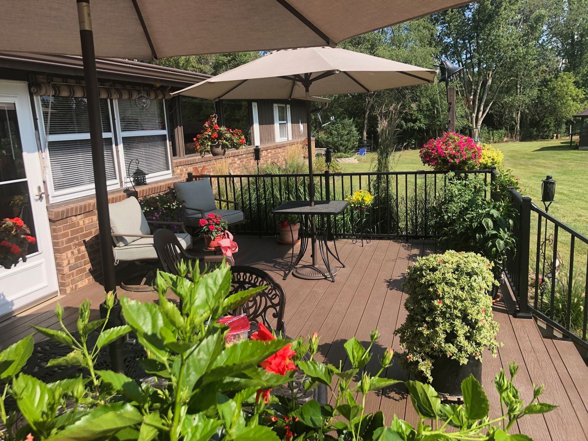 A brown backyard deck features patio furniture, two tan umbrellas, and potted flowers under a sunny sky.