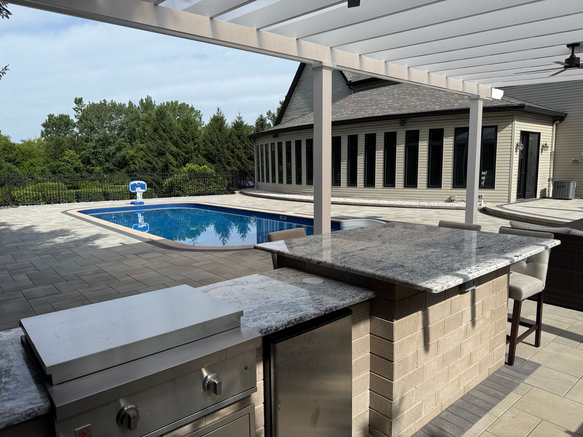 Outdoor kitchen with a grill and granite island under a pergola, facing a swimming pool and house on a sunny day.