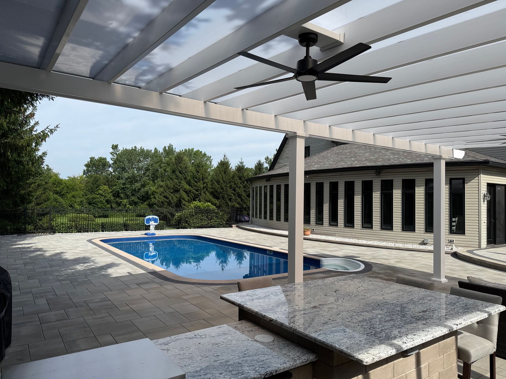 A covered patio with a granite countertop island and ceiling fan overlooks a backyard swimming pool and house.