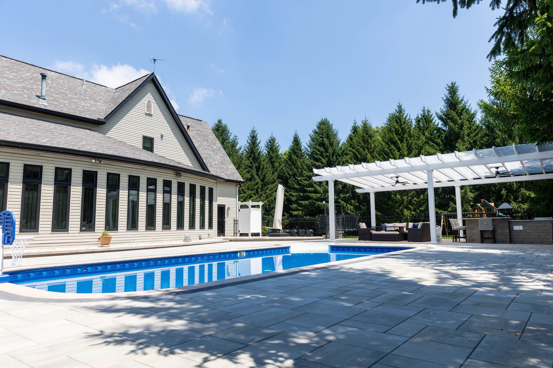 A sunny backyard scene featuring a swimming pool, a stone patio, and a white pergola with outdoor seating.