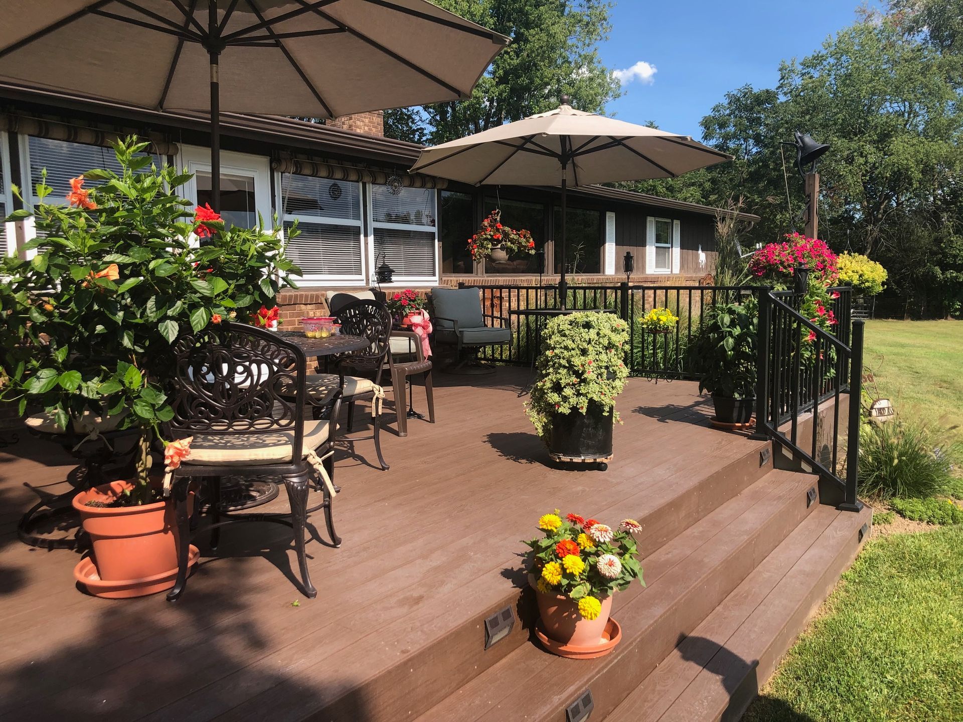 A brown wooden deck with patio furniture, umbrellas, and potted flowers under a sunny sky next to a house.