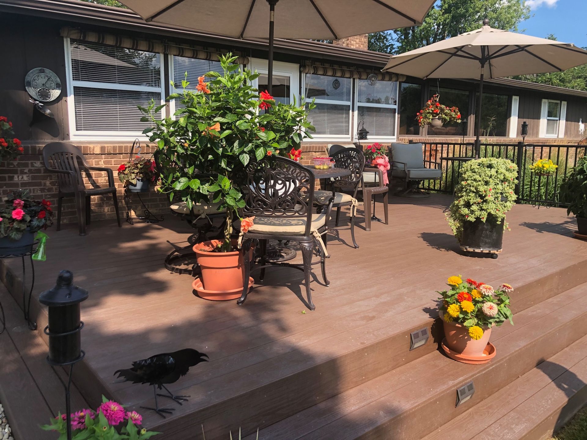 A sunny wooden deck with patio furniture, umbrellas, potted flowers, and a decorative black bird statue on the floor.