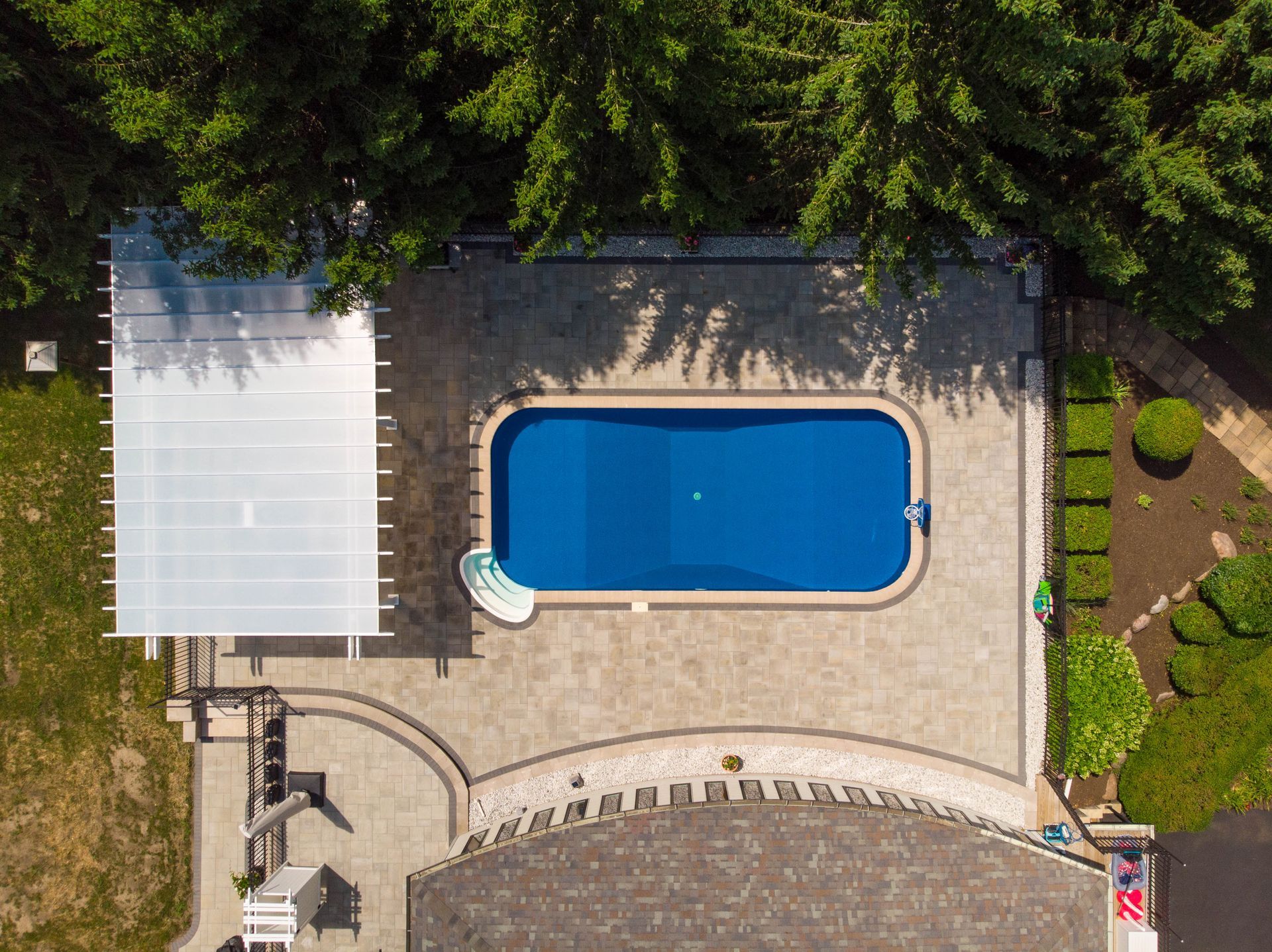 Aerial view of a backyard with a rectangular blue swimming pool, light stone patio, white pergola, and surrounding trees.