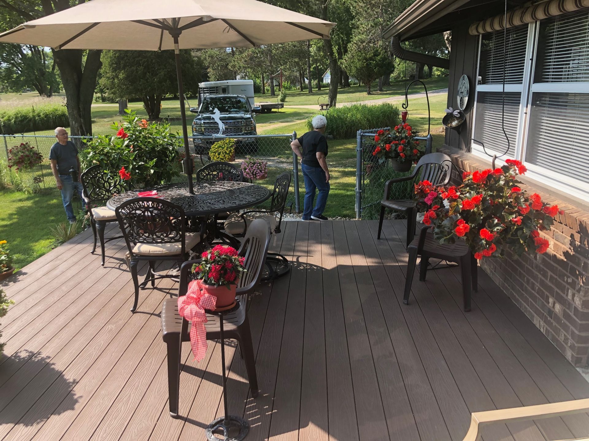 Two people stand on a residential wood deck featuring a round patio table, umbrella, chairs, and potted flowers.