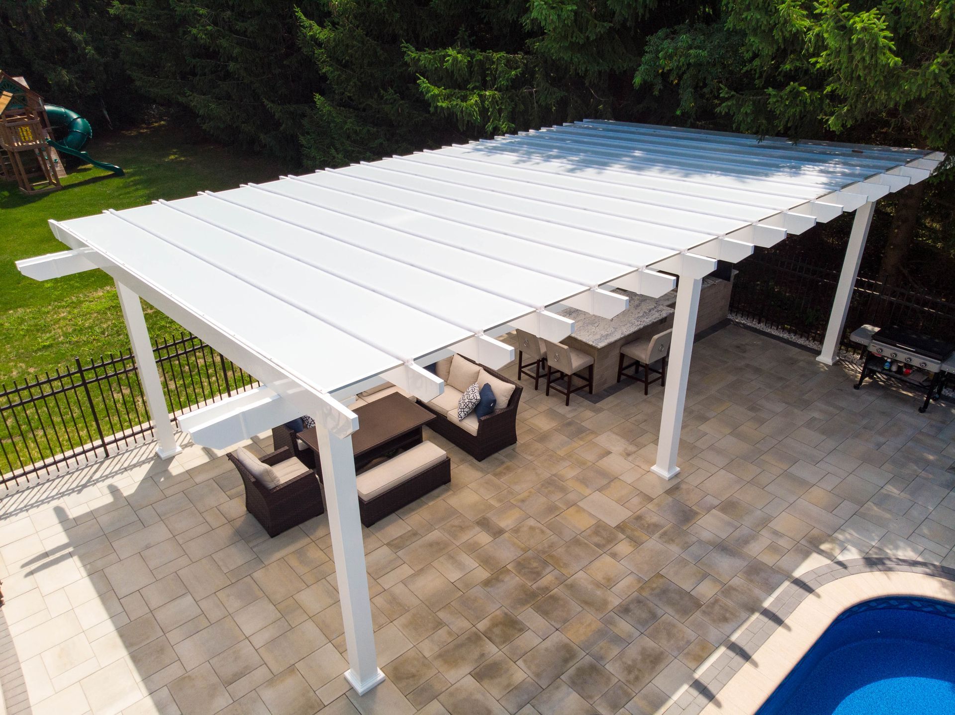 A white pergola provides shade over a paved patio featuring an outdoor dining set, a bar, and a nearby swimming pool.