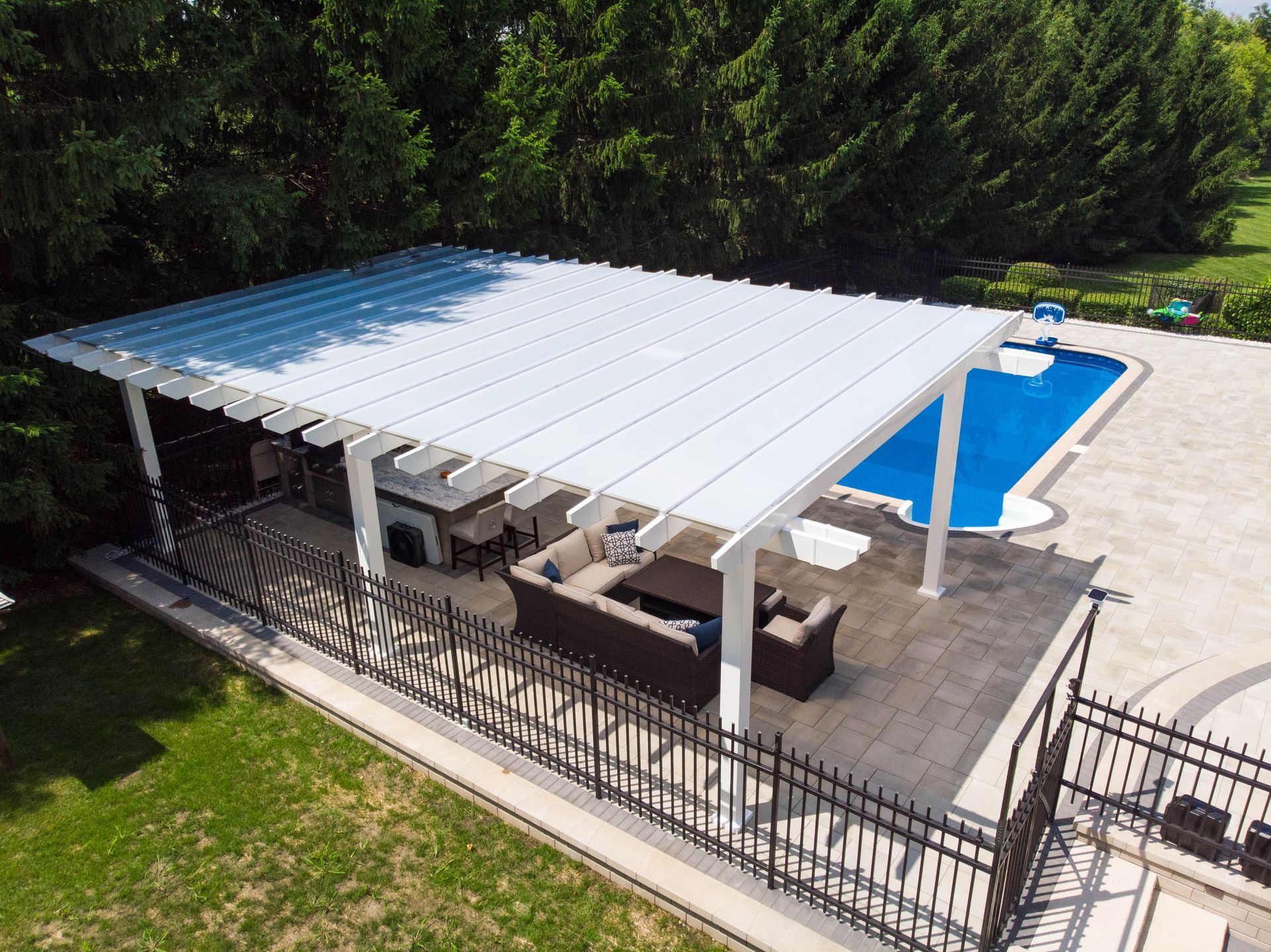 A white louvered pergola shades outdoor patio furniture next to a swimming pool in a residential backyard.