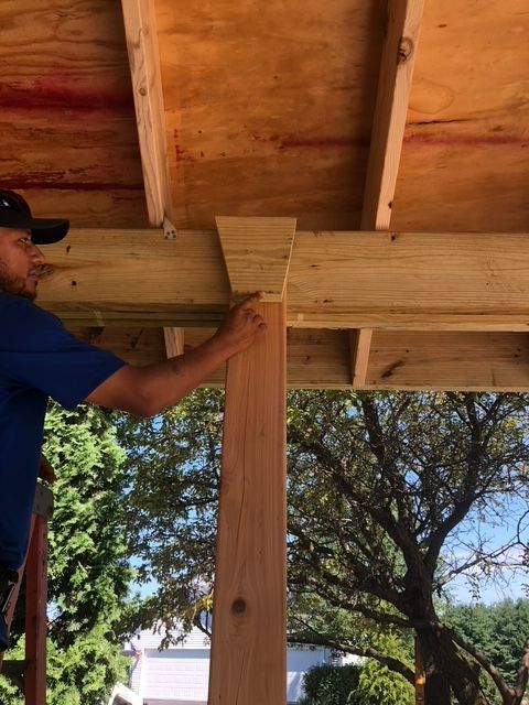 A worker points to a trapezoidal wooden support bracket connecting a vertical post to a horizontal beam on an outdoor roof.