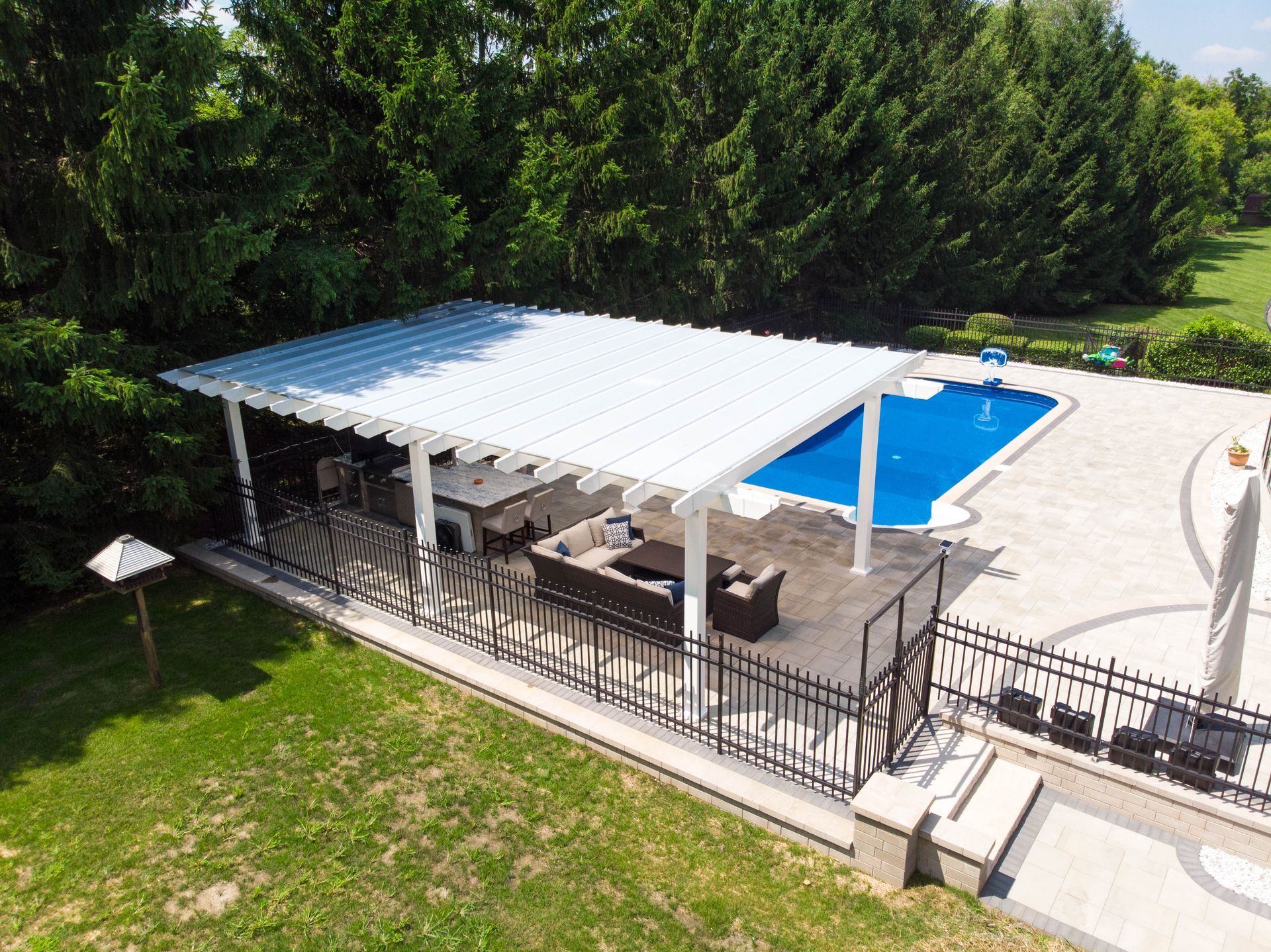 An aerial view of a white pergola with patio furniture next to a blue swimming pool in a lush backyard setting.